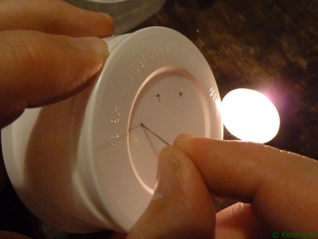 Mit einer heißen Nadel wird ein Loch in den Boden eines weißen Plastikgefäßes gestochen. Im Hintergrund brennt ein Teelicht.