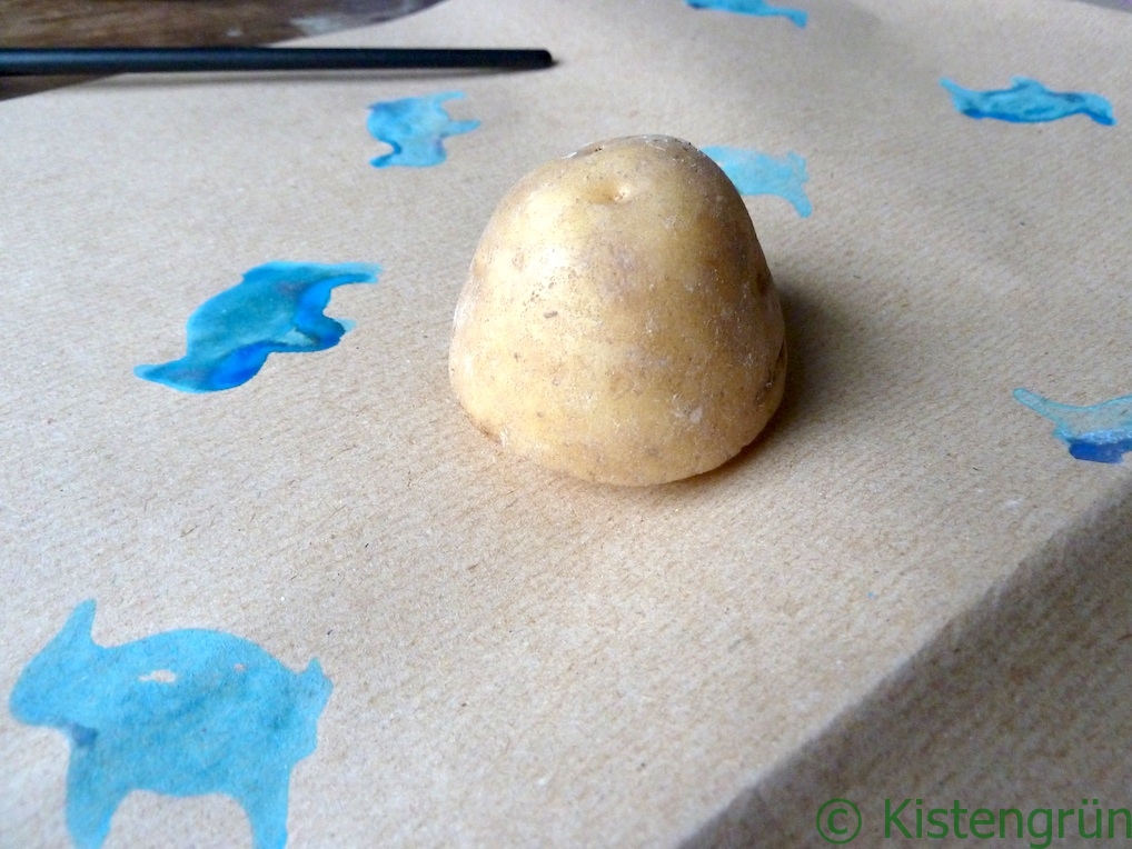 Kartoffeldruck: Eine hlabe Kartoffel liegt auf einem Packpapier, auf das schon blaue Pinguine gedruckt wurden.