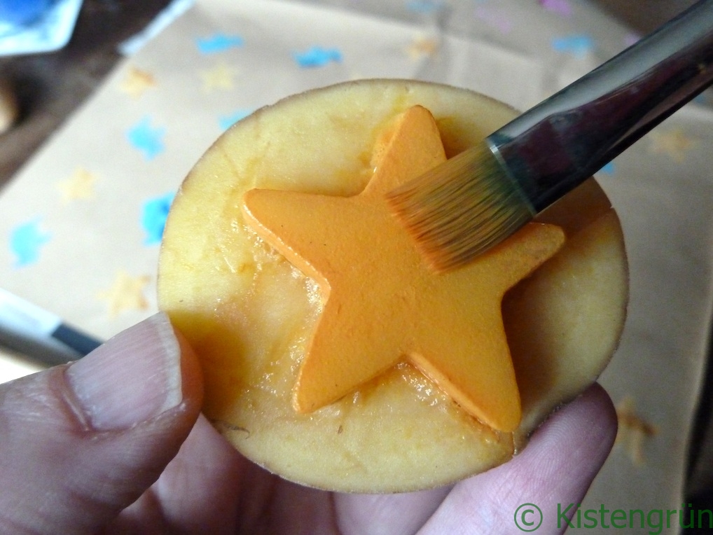 Auf einem Kartoffelstemepel wird ein Stern mit oragener Farbemit einem Pinsel bemalt.